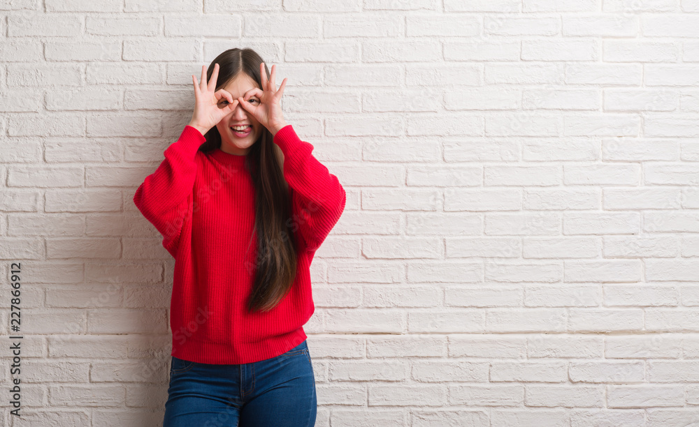 Young Chinese woman over brick wall doing ok gesture like binoculars sticking tongue out, eyes looking through fingers. Crazy expression. Young Chinese woman over brick wall doing ok gesture like binoculars sticking tongue out, eyes looking through fingers. Crazy expression.