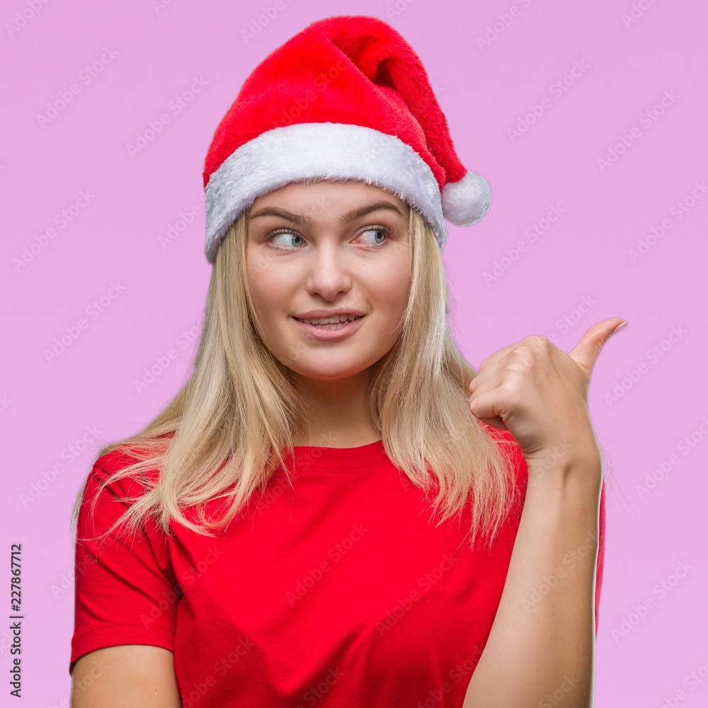 Young caucasian woman wearing christmas hat over isolated background smiling with happy face looking and pointing to the side with thumb up. Young caucasian woman wearing christmas hat over isolated background smiling with happy face looking and pointing to the side with thumb up.