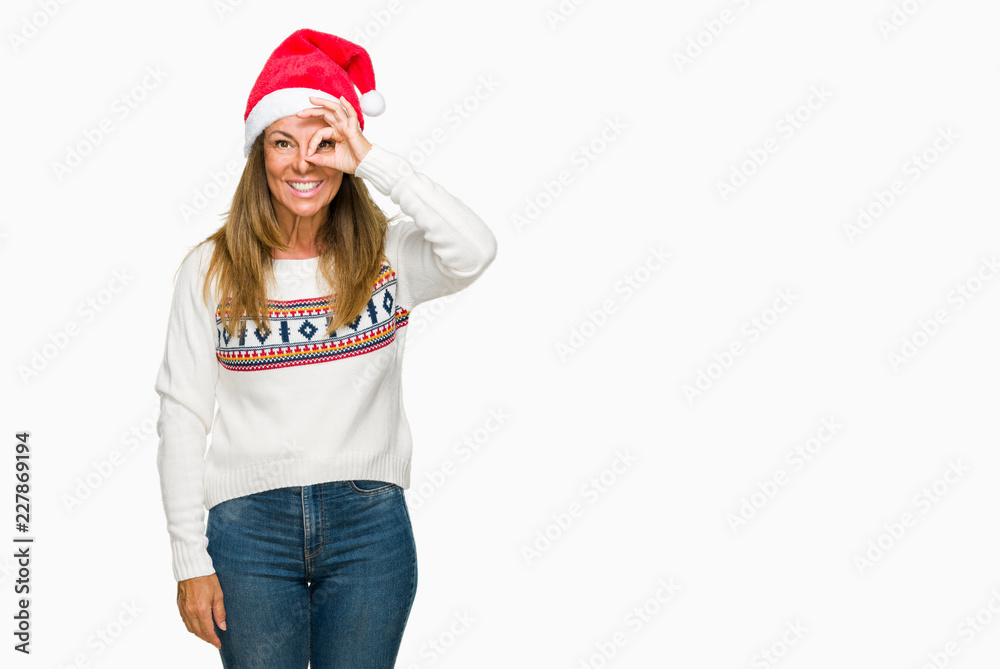 Middle age adult woman wearing winter sweater and chrismat hat over isolated background doing ok gesture with hand smiling, eye looking through fingers with happy face. Middle age adult woman wearing winter sweater and chrismat hat over isolated background doing ok gesture with hand smiling, eye looking through fingers with happy face.