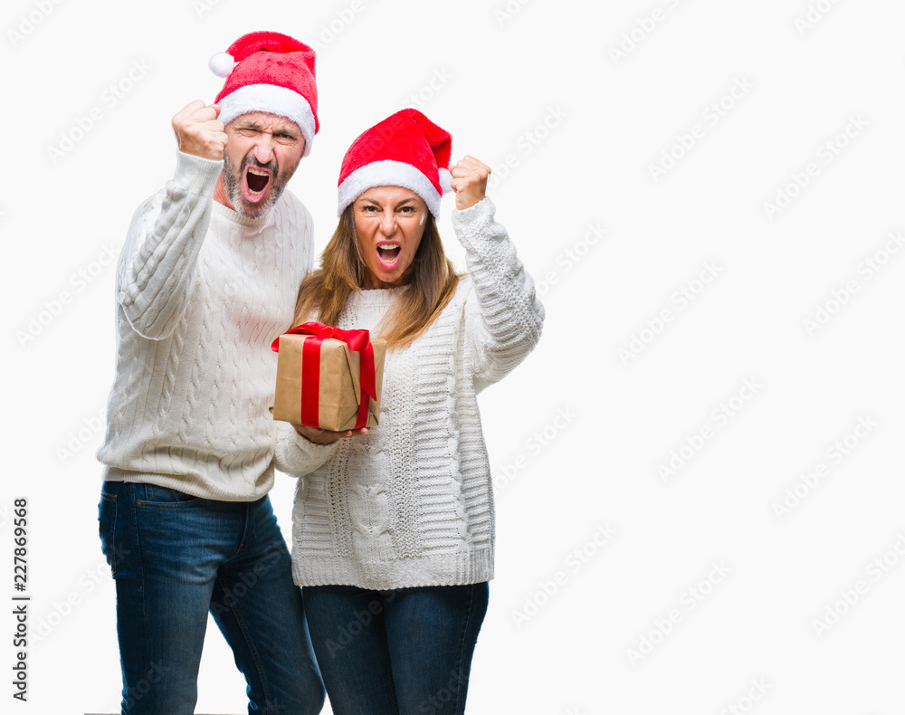 Middle age hispanic couple wearing christmas hat and holding gift over isolated background annoyed and frustrated shouting with anger, crazy and yelling with raised hand, anger concept Middle age hispanic couple wearing christmas hat and holding gift over isolated background annoyed and frustrated shouting with anger, crazy and yelling with raised hand, anger concept