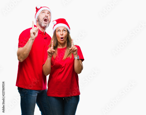 Middle age hispanic couple wearing christmas hat over isolated background amazed and surprised looking up and pointing with fingers and raised arms.