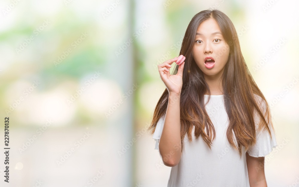 Young asian woman eating pink macaron sweet over isolated background scared in shock with a surprise face, afraid and excited with fear expression