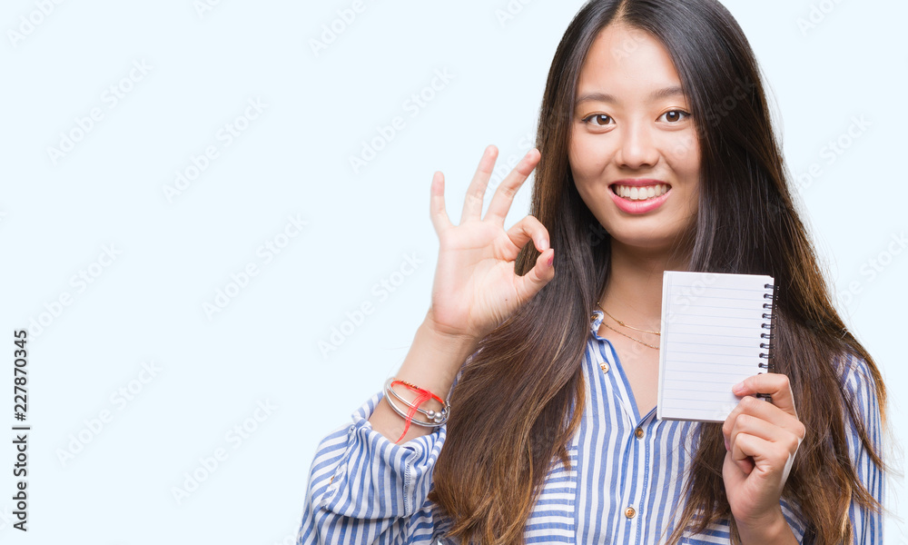 Young asian woman holding notebook over isolated background doing ok sign with fingers, excellent symbol Young asian woman holding notebook over isolated background doing ok sign with fingers, excellent symbol