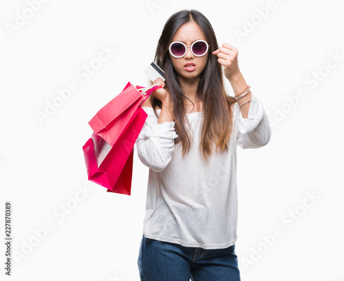Young asian woman holding shopping bags on sales over isolated background annoyed and frustrated shouting with anger, crazy and yelling with raised hand, anger concept