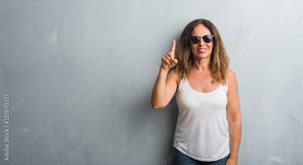 Middle age hispanic woman over grey wall wearing sunglasses showing and pointing up with finger number one while smiling confident and happy. Middle age hispanic woman over grey wall wearing sunglasses showing and pointing up with finger number one while smiling confident and happy.