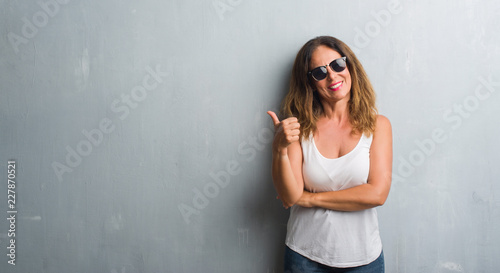 Middle age hispanic woman over grey wall wearing sunglasses smiling with happy face looking and pointing to the side with thumb up.