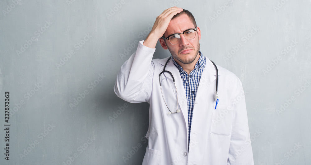 Young caucasian man over grey grunge wall wearing doctor uniform stressed with hand on head, shocked with shame and surprise face, angry and frustrated. Fear and upset for mistake. Young caucasian man over grey grunge wall wearing doctor uniform stressed with hand on head, shocked with shame and surprise face, angry and frustrated. Fear and upset for mistake.
