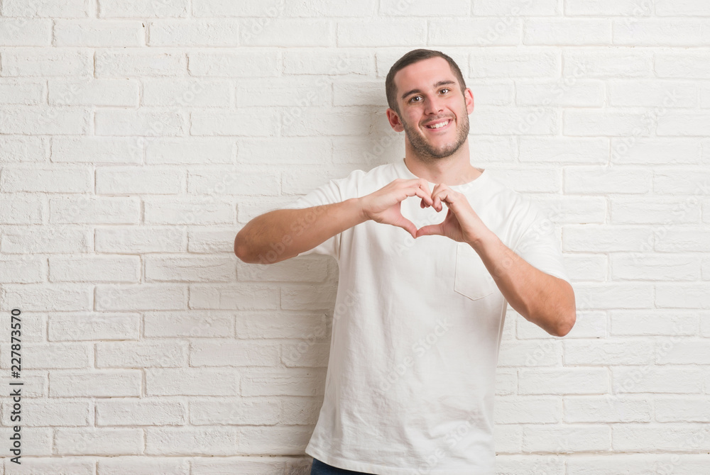 Young caucasian man standing over white brick wall smiling in love showing heart symbol and shape with hands. Romantic concept. Young caucasian man standing over white brick wall smiling in love showing heart symbol and shape with hands. Romantic concept.
