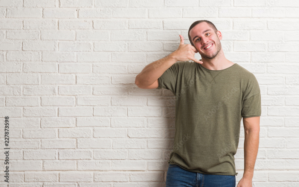 Young caucasian man standing over white brick wall smiling doing phone gesture with hand and fingers like talking on the telephone. Communicating concepts. Young caucasian man standing over white brick wall smiling doing phone gesture with hand and fingers like talking on the telephone. Communicating concepts.
