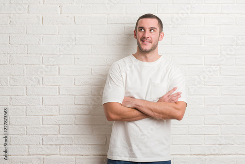 Young caucasian man standing over white brick wall smiling looking side and staring away thinking.