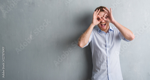 Young redhead business man over grey grunge wall doing ok gesture like binoculars sticking tongue out, eyes looking through fingers. Crazy expression.