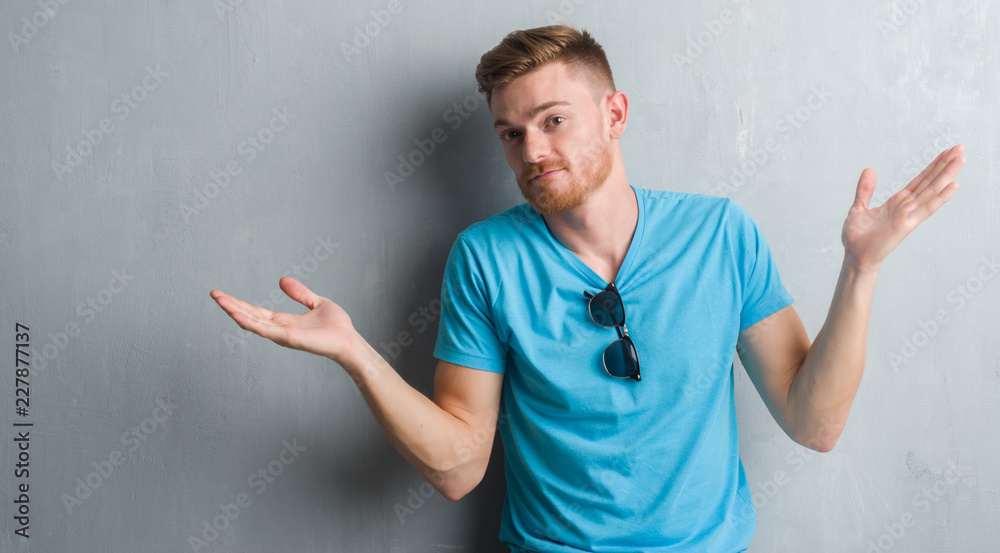 Fototapeta premium Young redhead man over grey grunge wall wearing casual outfit clueless and confused expression with arms and hands raised. Doubt concept.
