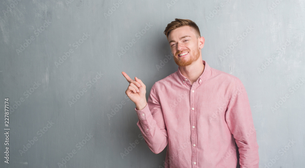 Young redhead man over grey grunge wall wearing pink shirt with a big smile on face, pointing with hand and finger to the side looking at the camera. Young redhead man over grey grunge wall wearing pink shirt with a big smile on face, pointing with hand and finger to the side looking at the camera.