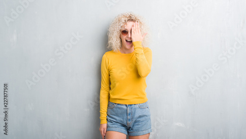 Young blonde woman with curly hair over grunge grey background covering one eye with hand with confident smile on face and surprise emotion.