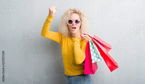 Young blonde woman over grunge grey background holding shopping bags on sales annoyed and frustrated shouting with anger, crazy and yelling with raised hand, anger concept