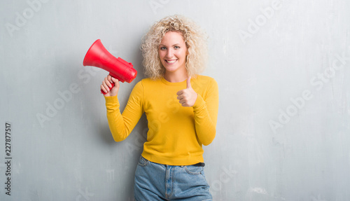 Young blonde woman over grunge grey background holding megaphone happy with big smile doing ok sign, thumb up with fingers, excellent sign