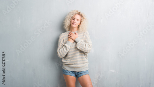 Young blonde woman with curly hair over grunge grey background smiling with hands on chest with closed eyes and grateful gesture on face. Health concept.