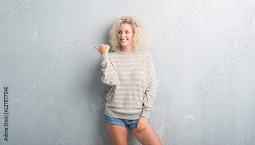 Young blonde woman with curly hair over grunge grey background smiling with happy face looking and pointing to the side with thumb up. © Krakenimages.com