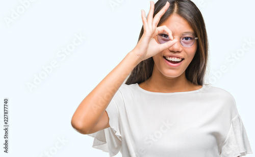 Young asian woman wearing glasses over isolated background doing ok gesture with hand smiling, eye looking through fingers with happy face. © Krakenimages.com