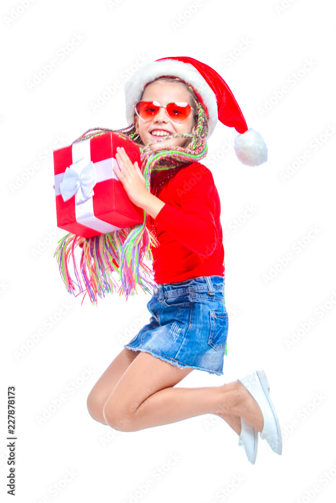 Obraz premium Girl in Santa's hat. Portrait of little cute girl holding box of Christmas present, Small girl hug her gift with happy and fun expression isolated on white background. Jump up. Holiday gift