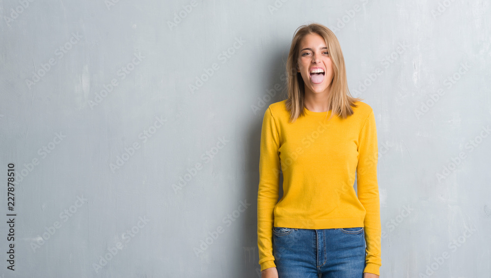 Beautiful young woman standing over grunge grey wall sticking tongue out happy with funny expression. Emotion concept. Beautiful young woman standing over grunge grey wall sticking tongue out happy with funny expression. Emotion concept.