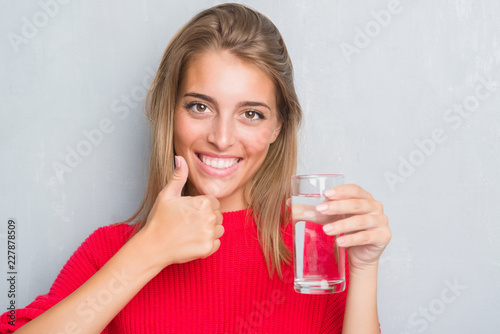 Beautiful young woman over grunge grey wall drinking a glass of water happy with big smile doing ok sign, thumb up with fingers, excellent sign