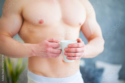 Sexy handsome young man standing shirtless in his bedroom, holding a coffee cup