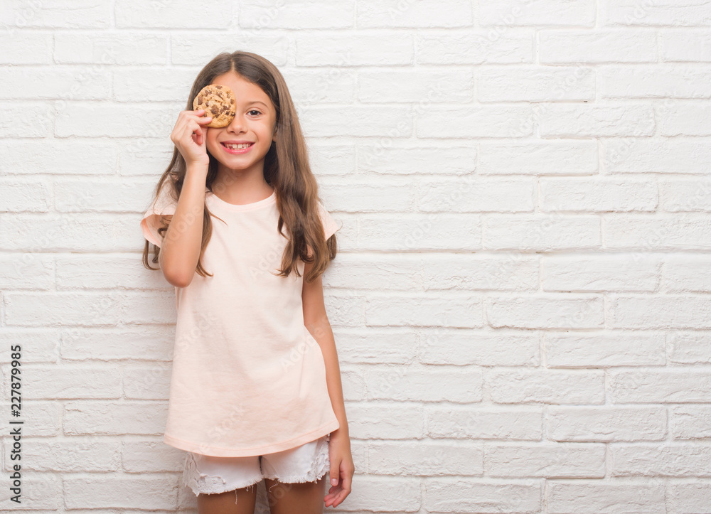 Young hispanic kid over white brick wall eating chocolate chips cooky with a happy face standing and smiling with a confident smile showing teeth Young hispanic kid over white brick wall eating chocolate chips cooky with a happy face standing and smiling with a confident smile showing teeth