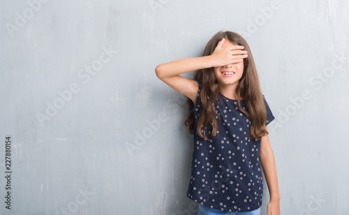 Young hispanic kid over grunge grey wall smiling and laughing with hand on face covering eyes for surprise. Blind concept.