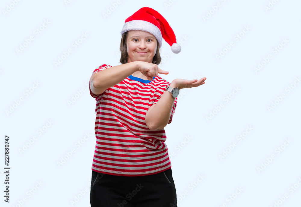 Young adult woman with down syndrome wearing christmas hat over isolated background amazed and smiling to the camera while presenting with hand and pointing with finger. Young adult woman with down syndrome wearing christmas hat over isolated background amazed and smiling to the camera while presenting with hand and pointing with finger.