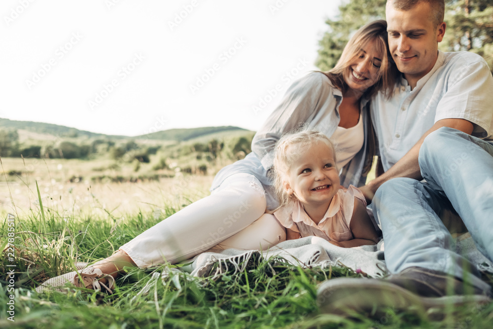 Fototapeta premium Young Adult Couple with Their Little Daughter Having Fun in the Park Outside the City, Family Weekend Picnic Concept, Three People Enjoying Summer Time
