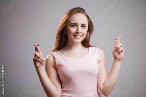 Beautiful young Caucasian woman wearing pink dress and with broad smile crosses fingers on grey background