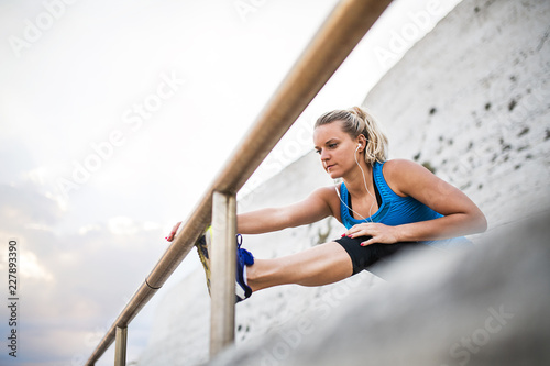 Young sporty woman runner with earphones stretching on the beach outside.