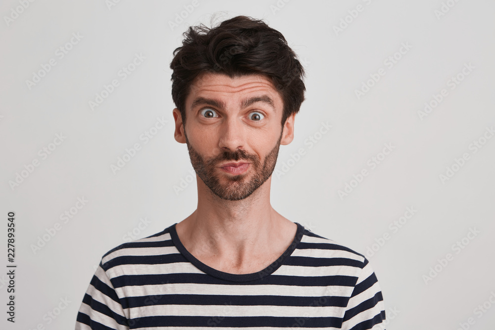 Clouse up of young brunet with beard looking suddenly surprised and discontent, eyebrows raised, lips pursed wears black and white striped tshirt, stands rightside isolated over white background Clouse up of young brunet with beard looking suddenly surprised and discontent, eyebrows raised, lips pursed wears black and white striped tshirt, stands rightside isolated over white background
