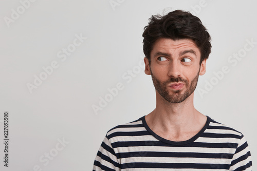 Clouse up of young brunet with beard looking to the left with distrust and discontent, frowning brows, lips pursed wears black and white striped tshirt, stands rightside isolated over white background