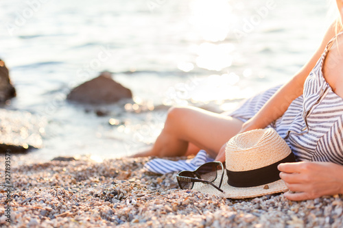 Woman is laying on sea beach at sunset. Girl is wearing in casual striped dress with straw hat, sunglasses. Beautiful female traveler is relaxing, enjoying life in summer vacation.
