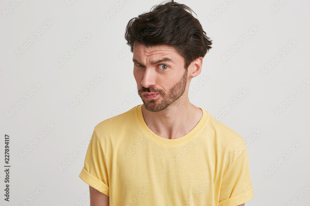 Dark hair young man with beard wearing yellow tshirt looks suspicious and showing defiance,isolated over white background Dark hair young man with beard wearing yellow tshirt looks suspicious and showing defiance,isolated over white background