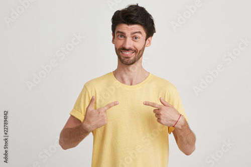 Smiling young man with dark brown hair and beard, shows with fingers at himself, wearing yellow tshirt, isolated over white background