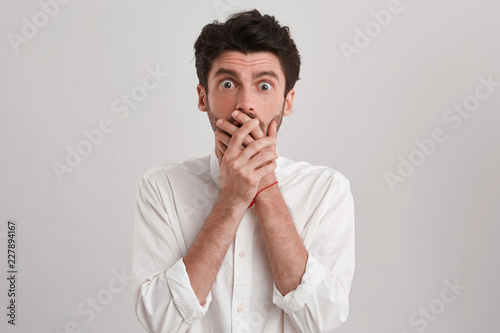 Portrait of young man wearing white shirt looks astonished shocked and frightened  mouth closed with both palms  isolated over white background