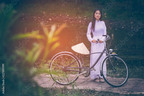 Portrait of beautiful young Asian teen with long hair wearing a costume of Viet Nam (Ao dai) she is currently towed a bicycle and holding a hat by verdant gardens background with copy space.