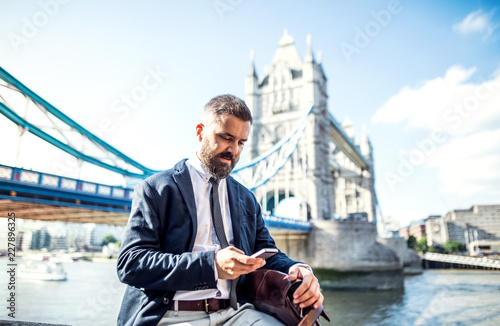Photography Hipster businessman with smartphone sitting by the Tower Bridge in London