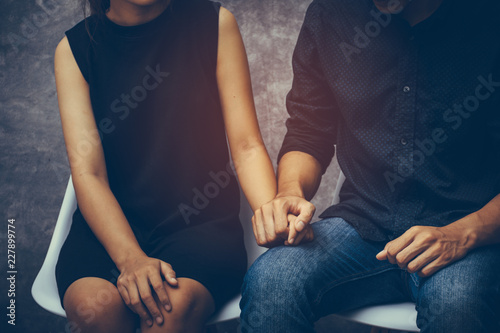 Young couples sitting on a chair inside the room which walls are made from cement. They shake hands, show their love and encouragement toward each other with copy space.