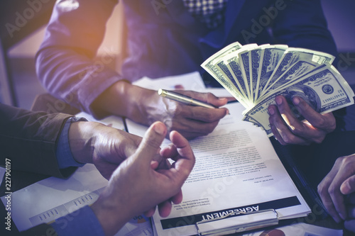 Closeup picture stack of banknotes US dollars in the hands of young men. In his right hand holding a pen and signing documents on business deals by having a third party become a witness.