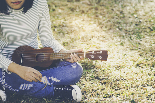 Asian teen girl wearing jeans and T-shirt playing ukulele is within the park. She was sitting on the lawn in the warm sunlight shines from the back with copy space.