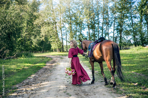 A woman with a bouquet of flowers dressed in a long burgundy dress with sleeves leads a brown horse along a rural road. The girl turns around. © Eno1