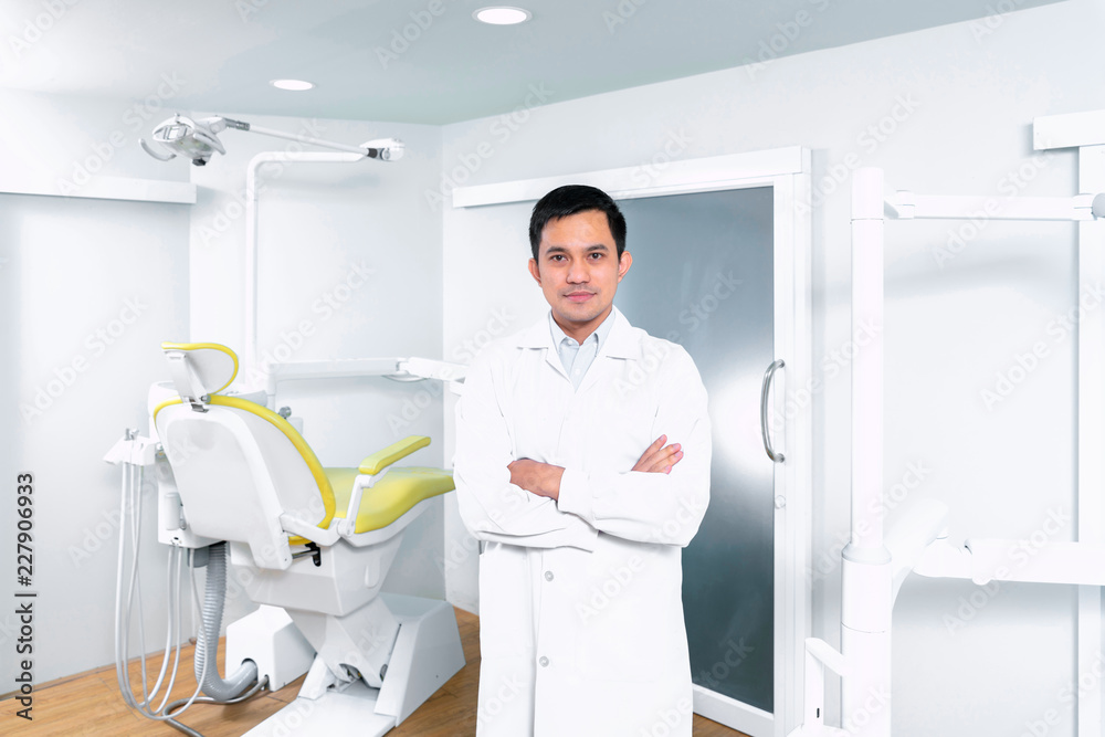 Cheerful Asian dentist standing in his clinic with his dental chair