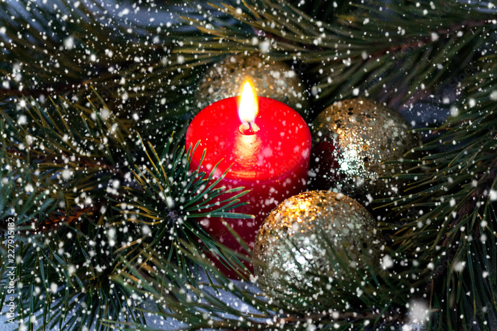 red burning candle and golden Christmas balls with a fir branch under going snow