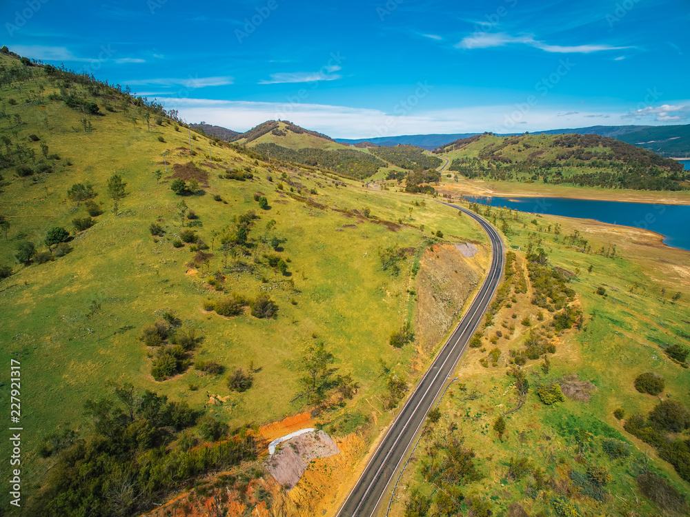 Fototapeta premium Rural road passing through green hills and lake - scenic aerial landscape