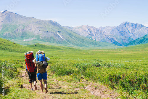 Travelers with large backpacks go on footpath in green valley to wonderful giant mountains with snow. Hiking in highlands. Amazing vivid mountain landscape of majestic nature. People climb on high.