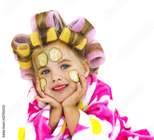 Девочка в банном розовом халате с бигудями и кусочками огурцов на лице.